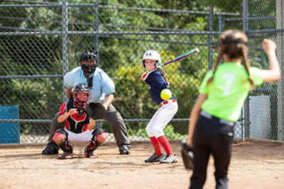 An image of a girl swinging a softball bat with a catcher and umpire behind her.