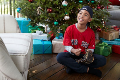 Boy in a red sox jersey and hat sits in front of a Christmas trees with presents underneath and smiles as he tries on his new glove.