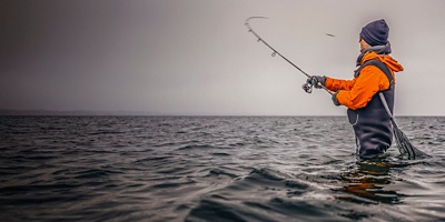 Angler in Wathose mit Spinnrute und Kescher beim Wurf mit Köder im Winter im kalten Meer an der Küste bei Wolken, Angler in Wathose mit Spinnrute und Kescher beim Wurf mit Köder