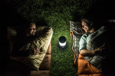 two girls smiling laying in sleeping bags in the grass at night with a lantern lit up between them