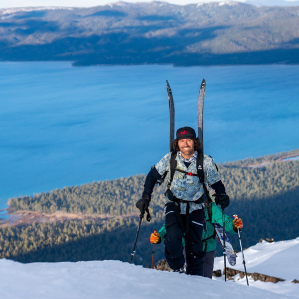 Jeremy Jones smiles for the camera hiking up a mountain with skis