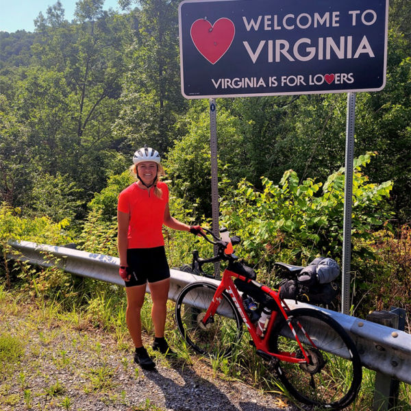 Brooke Barney poses with her bike in front of a Welcome to Virgina sign.