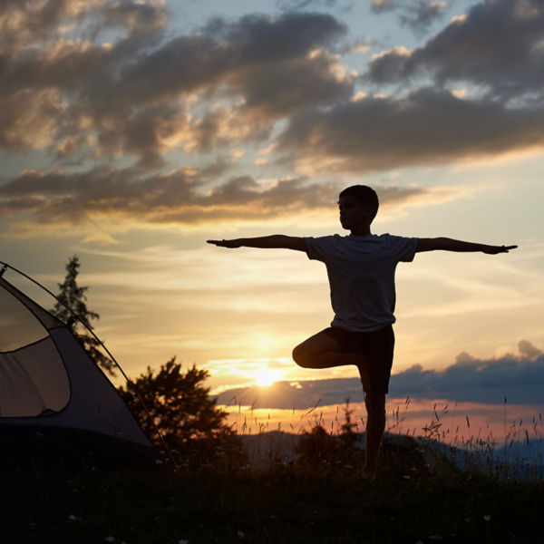 Silhouette of boy at practice yoga on the top of mountain at the dawn near campsite under sky with clouds and bright morning sun.