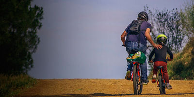 Father and son cycling up a hill. The father is helping his son by pushing him up the hill.