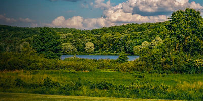 Landscape vista in Moraine State Park, in Southwest Pennsylvania in the evening.  Fields of green with Lake Arthur surrounded by trees and a bright blue cloud filled sky for a background.