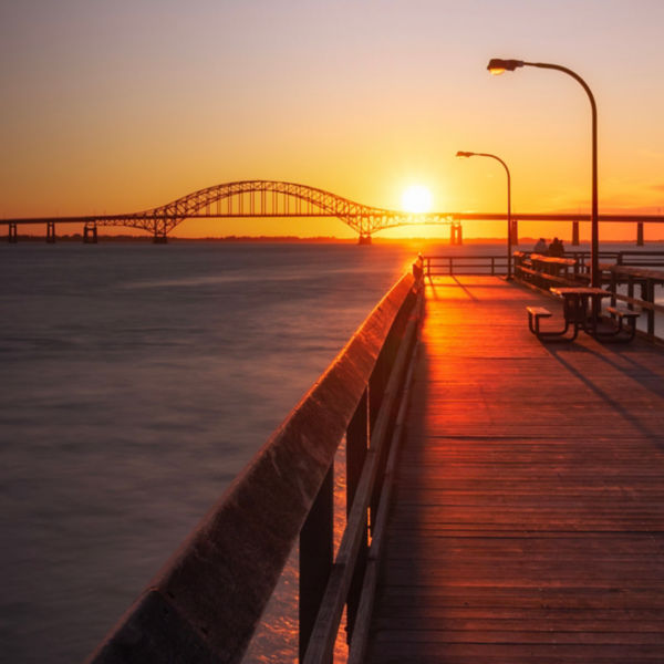 Wooden fishing pier leading towards an arched bridge, as the sunset dips below the horizon. Captree State Park - Long Island New York