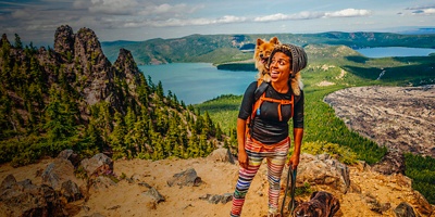 A woman hikes with her dog in a backpack and on a leash