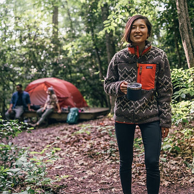 A woman standing and smiling while holding a mug of coffee, she is wearing a fleece multi-color quarter zip and black paints while standing in the forest.