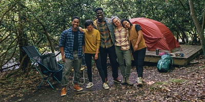 A group of five people smiling taking a picture in the forest with a tent and camp chairs around them.