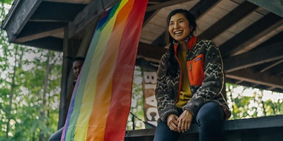 A woman sitting on a table next to a pride flag in a pavilion in the woods.