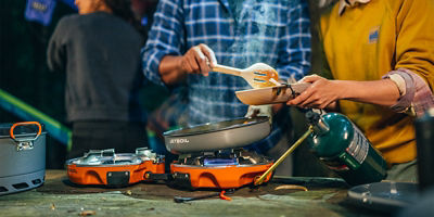 A person wearing a blue plaid flannel and another person wearing a mustard crewneck holding cookware while cooking using an outdoor table grill.