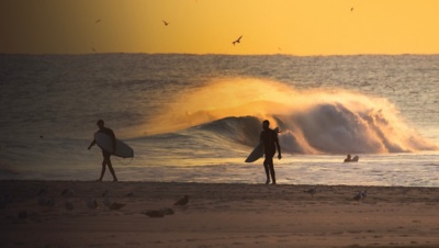Silhouette of a surfers walking on the beach at sunset, with waves crashing in the background. Long Beach New York