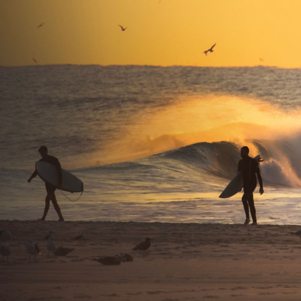 Silhouette of a surfers walking on the beach at sunset, with waves crashing in the background. Long Beach New York