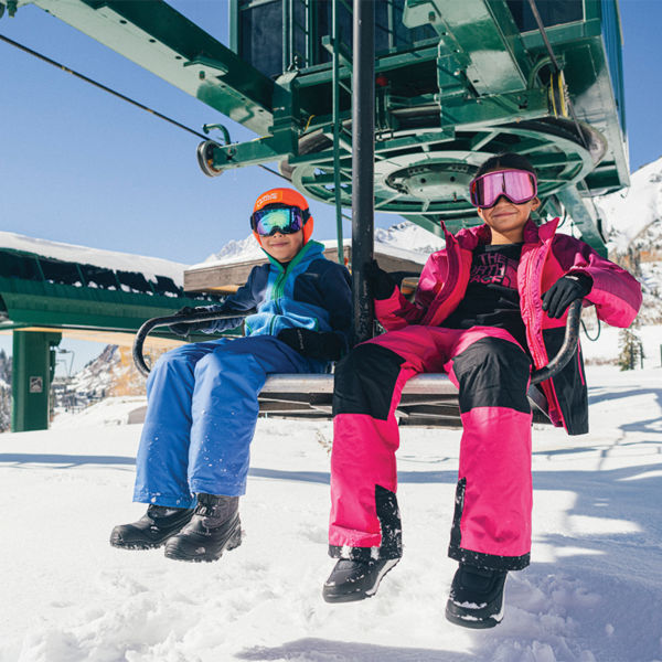 Two children sit on a ski lift in their ski and snowboard gear