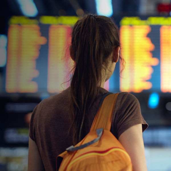 Young woman with small backpack in international airport looking at the flight information board, checking her flight