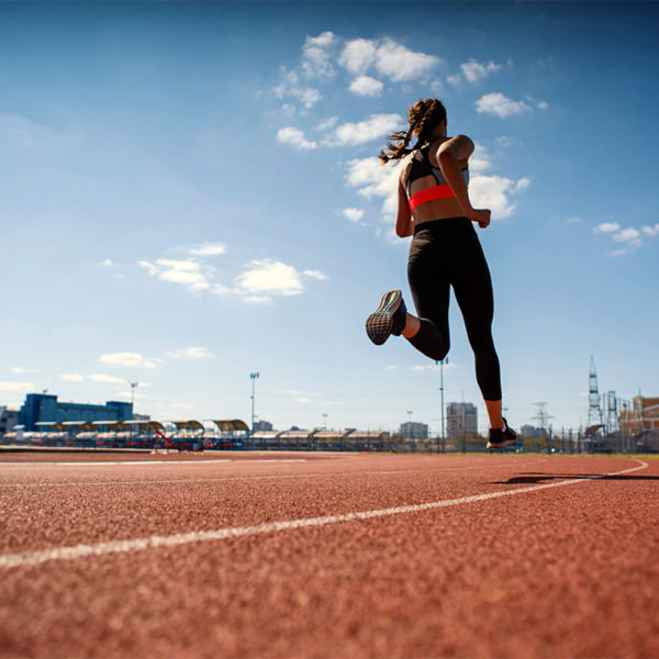 From below back view of teen firl in sportswear sprinting on running track at stadium under blue sky on summer day