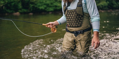 A man standing, wearing waders, while fishing in a creek.
