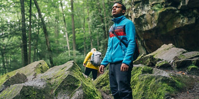 A man wearing a light blue full zip fleece and navy blue fleece joggers, standing between rocks in the forest.