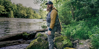 A man standing along a river side in the woods, holding a fishin rod and wearing a army green puffer jacket and grey pants.