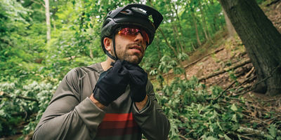 A man strapping on a bike helmet who is wearing amber sungglasses, grey long sleeve shirt and bike gloves while in the forest.