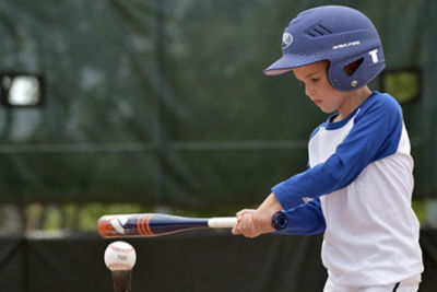 Little kid in a baseball shirt and batting helmet swings a baseball bat at a baseball sitting on a tee.
