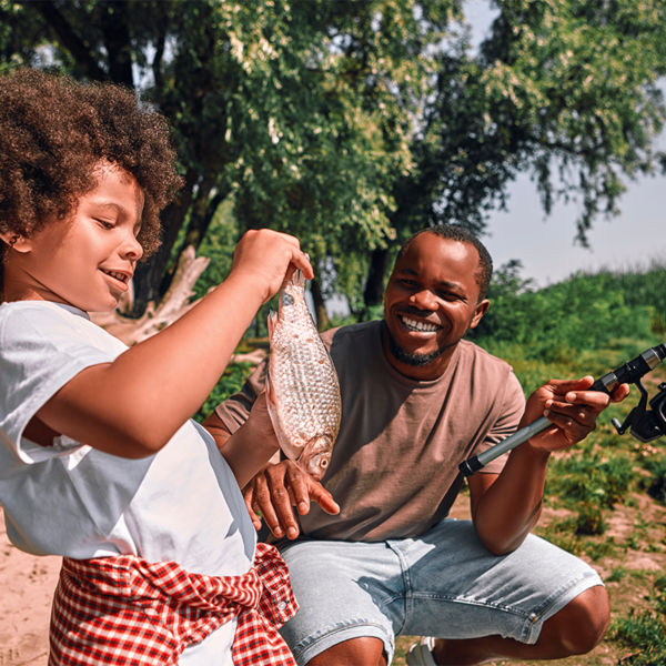 A boy looking at the fish in his hands while his dad holding a fishing rod