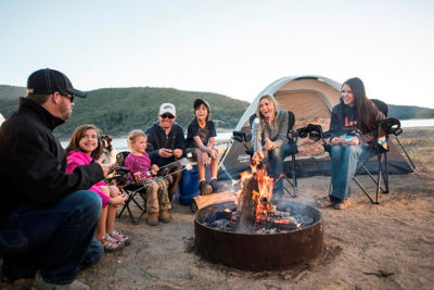 two families sitting around a campfire making smores with a lake in the background.