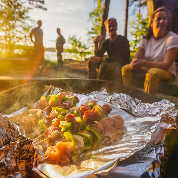 A group of people gather around food on skewers being grilled In a firepit 