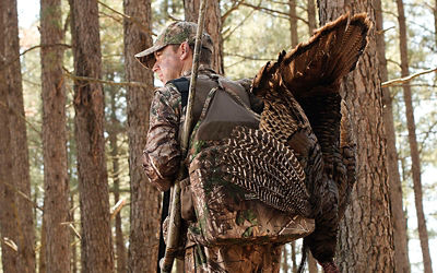 Picture of a man wearing camoflage and carrying a turkey while in the woods.