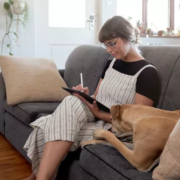 Young woman works from home on tablet with golden dog next to her on a couch.