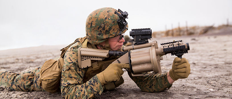 Marine aims M32 grenade launcher with mounted optics in prone position on beach during amphibious training exercise.
