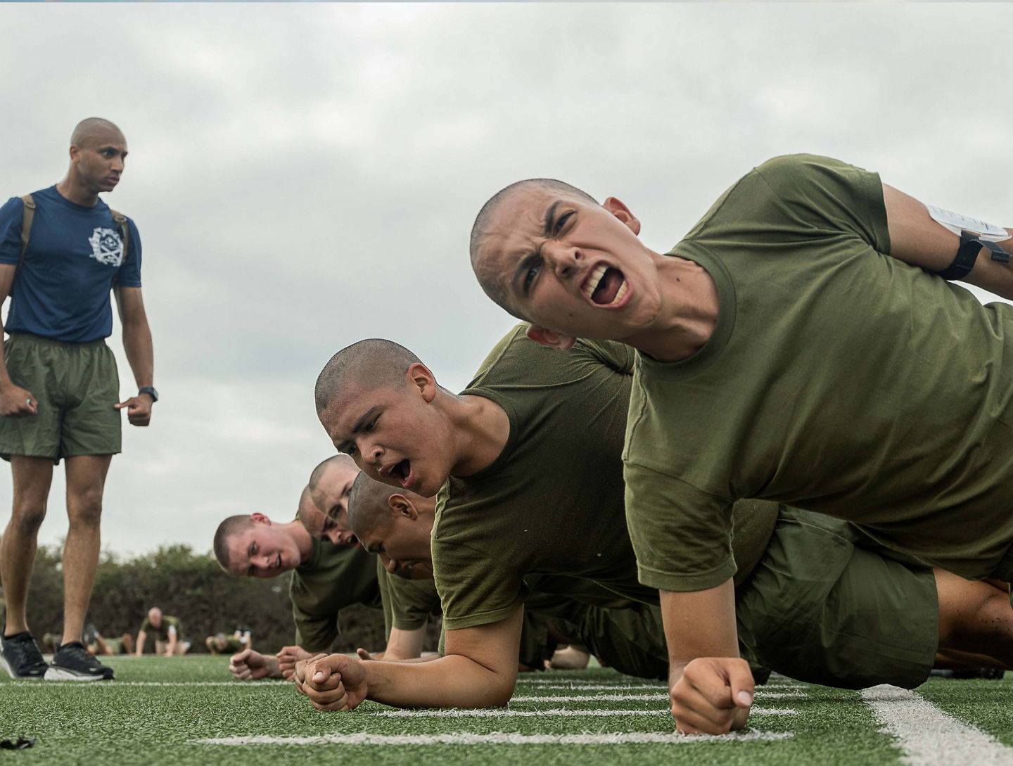 Marine Recruits train on a field. They are doing side planks and shouting while a drill instructor looks over them.