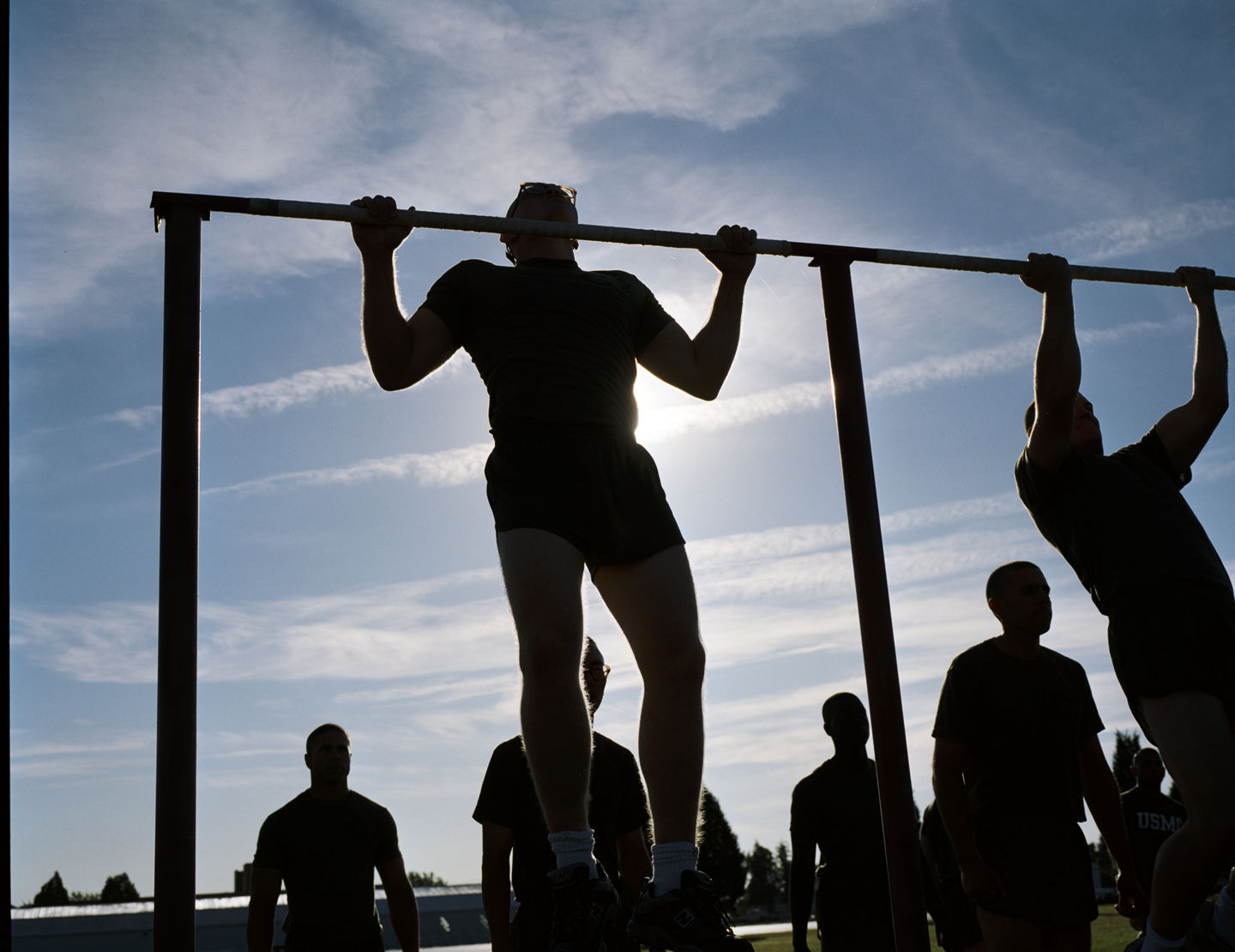 A marine recruit completes a pull-up during boot camp. He is backlit by the sun. You see his silhouette and the silhouettes of others. 