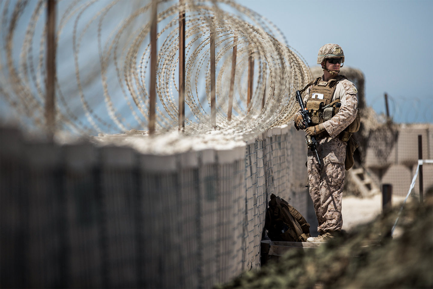 Marine sentry in desert camouflage with rifle standing guard at military checkpoint with security barriers and concertina wire.