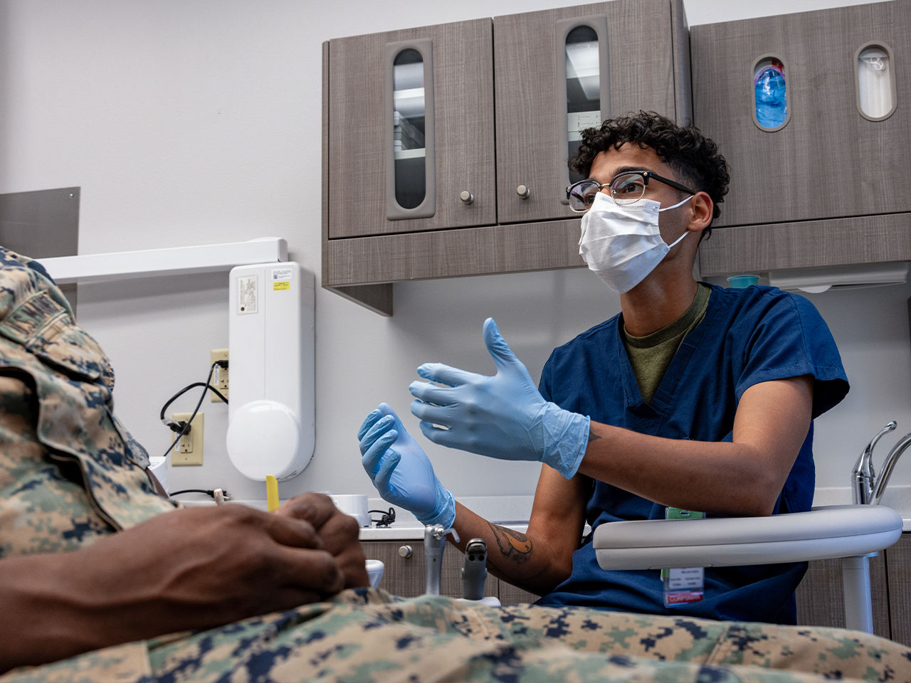 Military health professional in blue scrubs and mask providing medical care to Marine in clinical setting.
