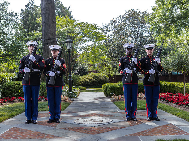 Four Marines in dress blues performing ceremonial rifle guard at memorial pathway outdoors.