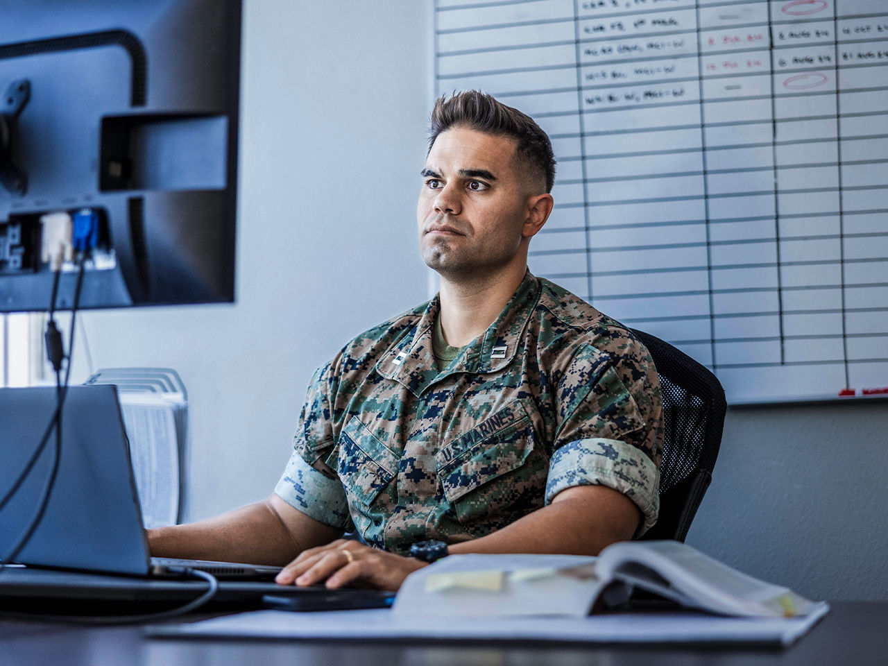 Marine Officer in camouflage uniform working at desk with computer and scheduling board in office setting.