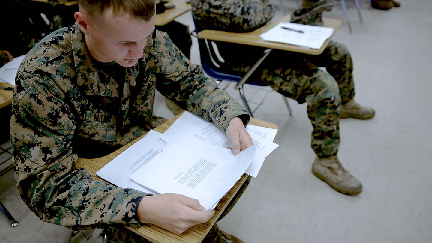 Marines in camouflage uniforms at a desk in a classroom setting.