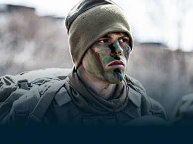 Marines in camouflage face paint and gear march in formation carrying packs during training exercise.