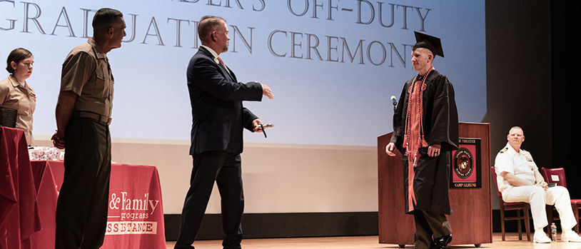Marine in dress uniform receiving diploma at graduation ceremony from university official on stage.