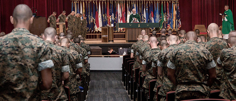 Marines attending religious service in chapel with chaplain conducting worship ceremony and state flags displayed.