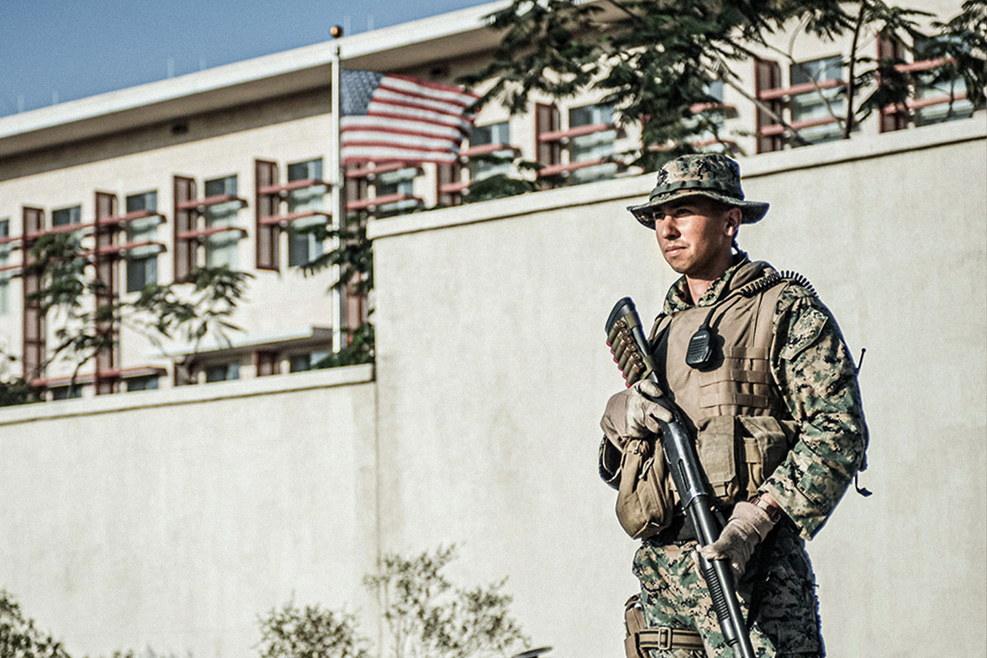 Marine in digital camouflage with tactical vest and rifle standing guard at embassy compound with American flag.