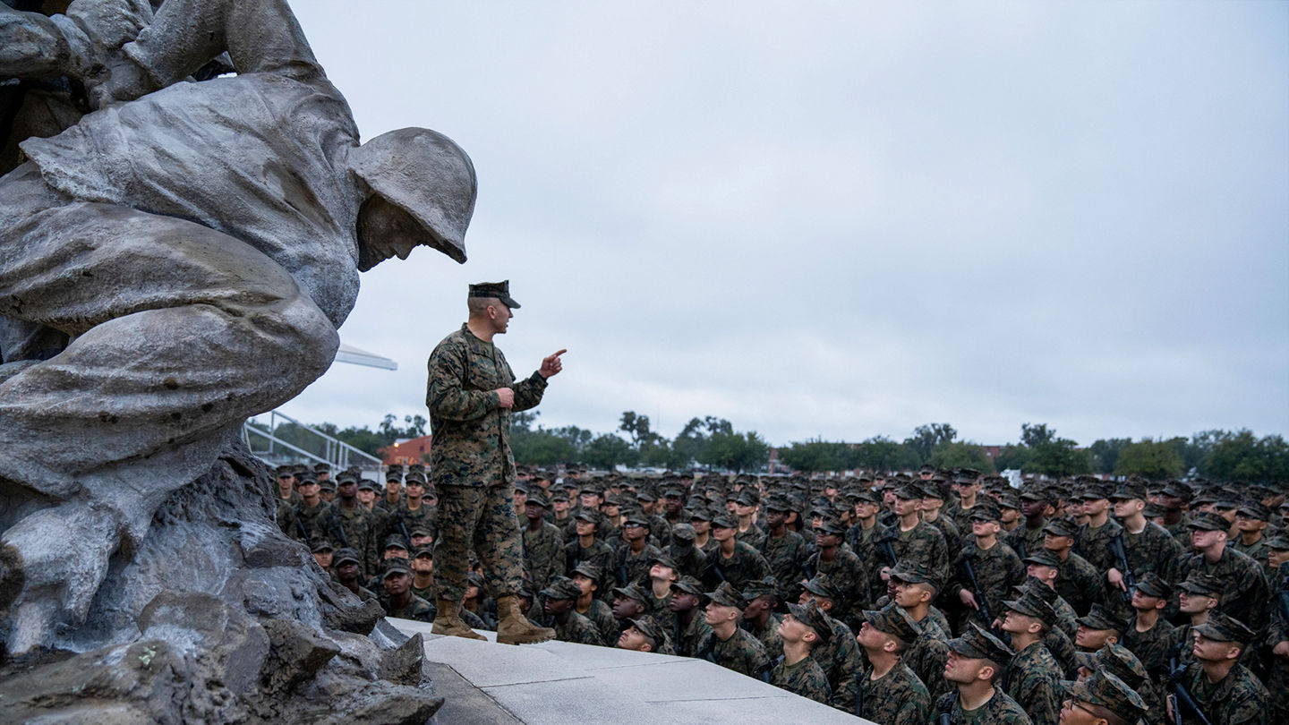 Marine leader addressing large formation of Marines gathered beside Iwo Jima Memorial during ceremony.