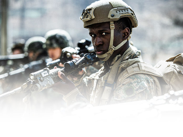 Marine in tan tactical helmet with optics-equipped rifle during precision shooting training exercise with team members.