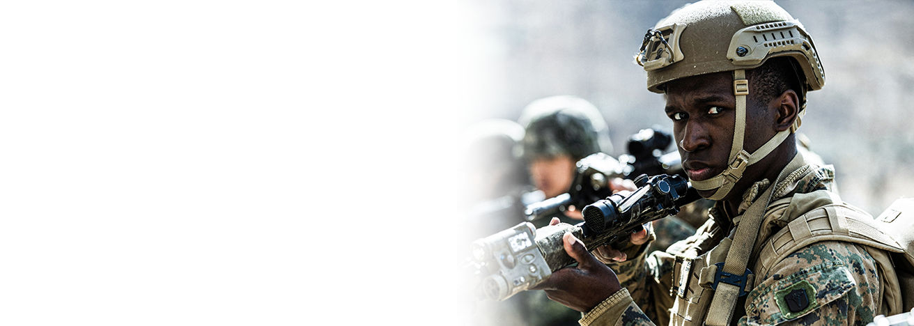 Marine in tan tactical helmet with optics-equipped rifle during precision shooting training exercise with team members.
