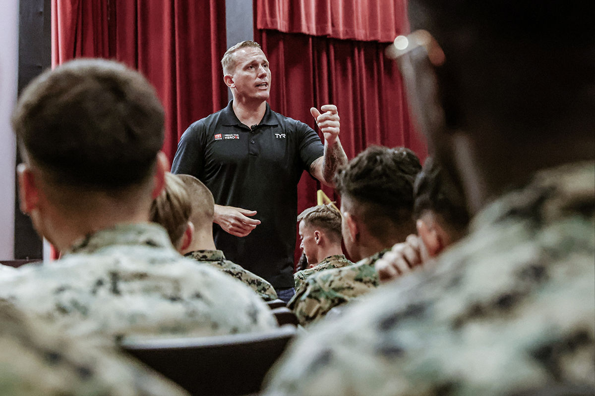 Speaker in black polo shirt addressing audience of Marines in woodland camouflage during motivational presentation or training session.
