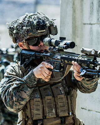 Marine marksman with scoped rifle and tactical gear conducting precision shooting training in urban environment.