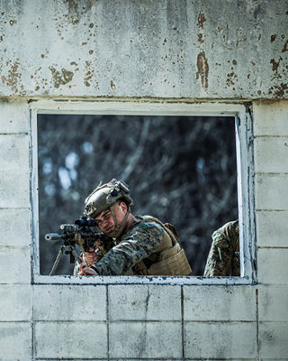 Marine sniper positioned at window with scoped rifle conducting urban combat training in South Korea.