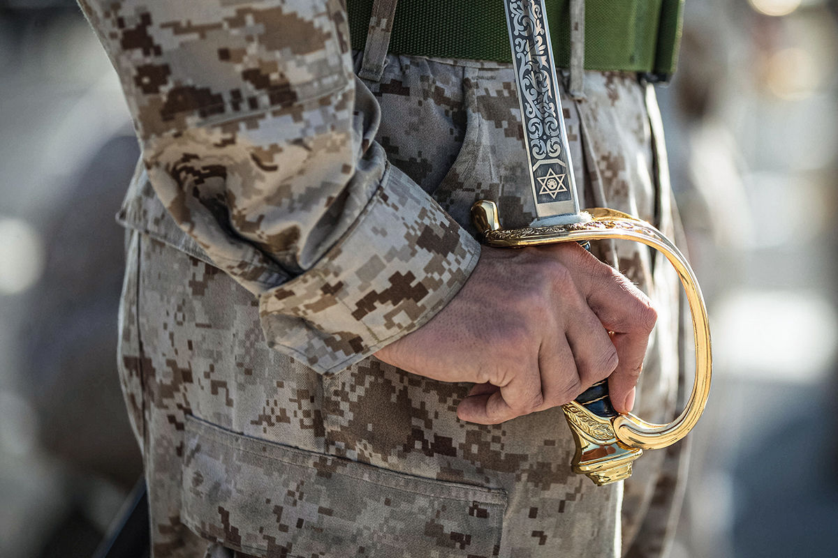 Close-up of NCO sword with ornate brass guard and engraved blade held by Marine in digital camouflage uniform.
