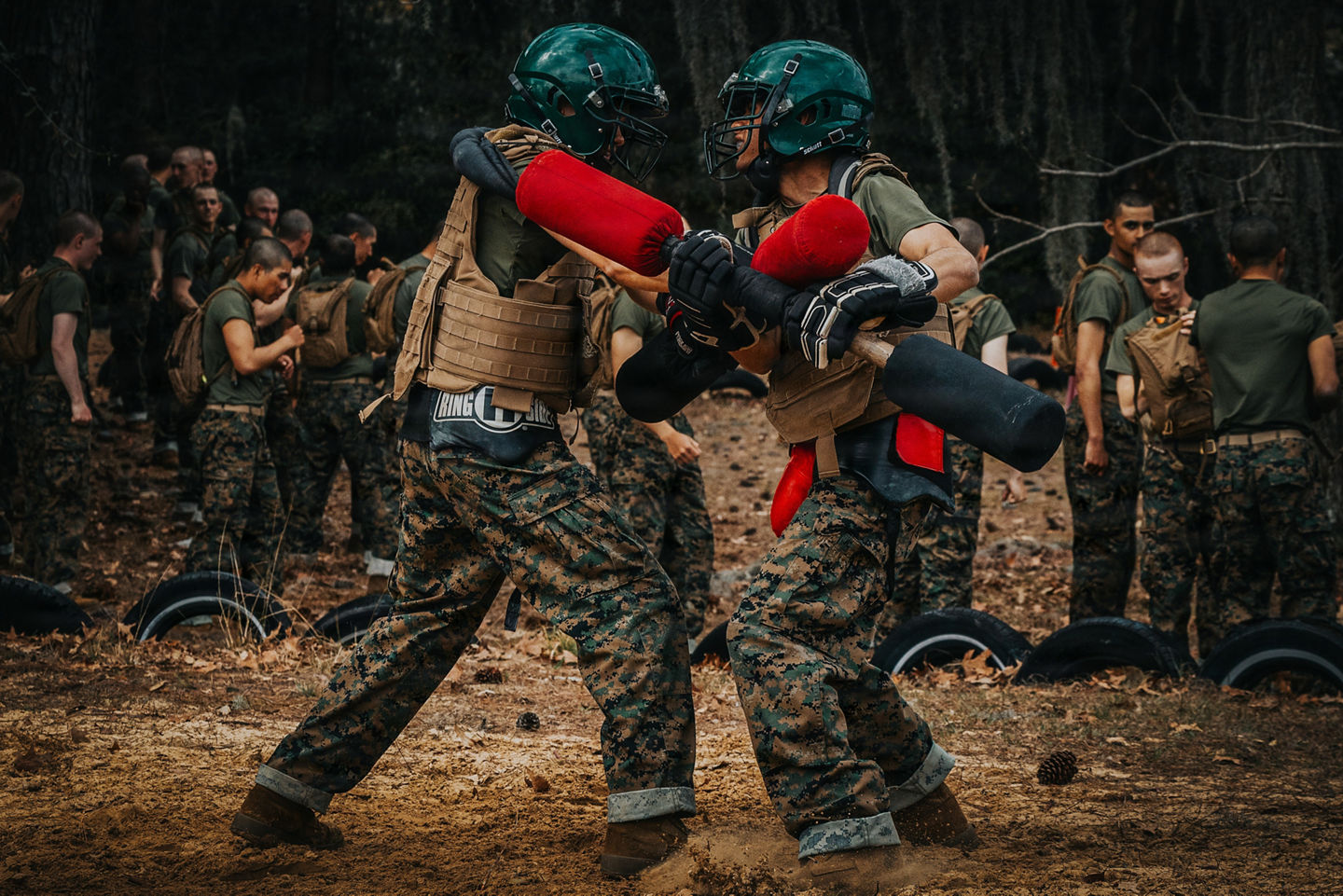Two Marine Corps recruits in helmets and protective gear spar with pugil sticks as other recruits look on at a wooded training pit.
